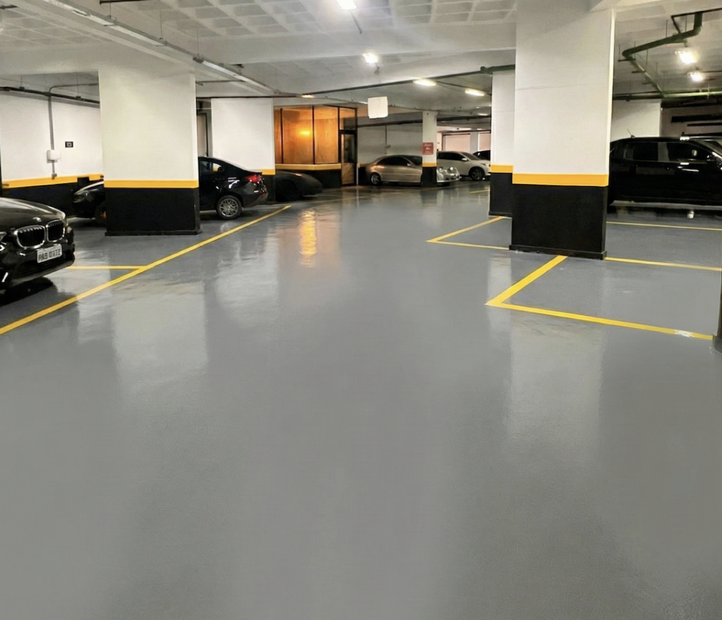 Piso epóxi aplicado em garagem de condomínio em Belo Horizonte, com acabamento cinza uniforme e sinalização viária amarela para vagas e circulação.